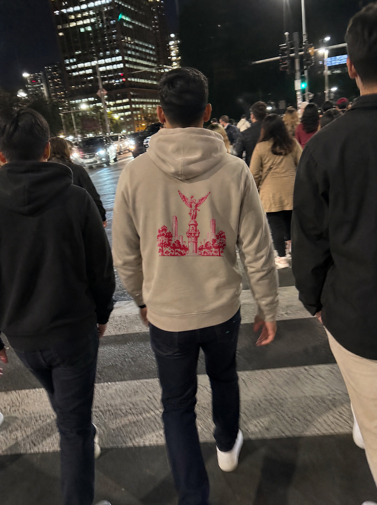 Man in a Mushroom El Ángel Hoodie, back view, crossing a busy downtown street at night with friends