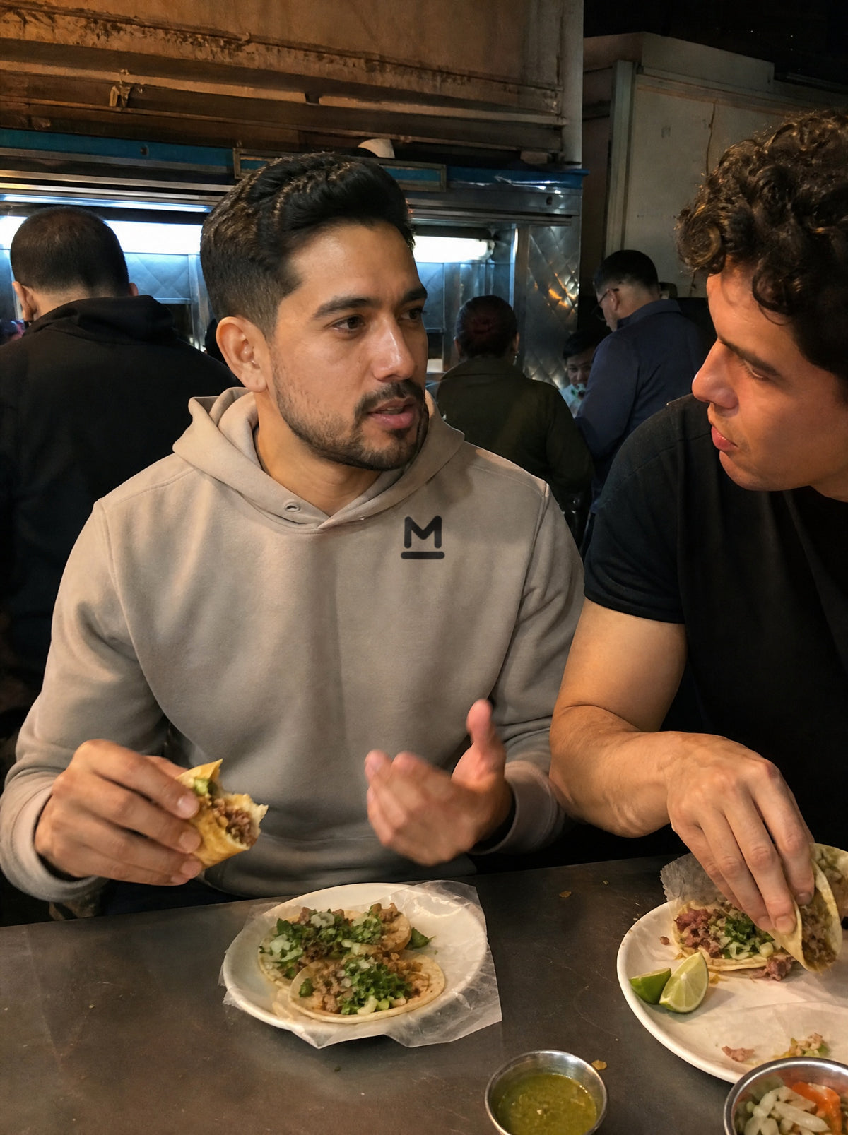 Man in a Mushroom El Ángel Hoodie eating tacos at a late night taquería counter with a friend
