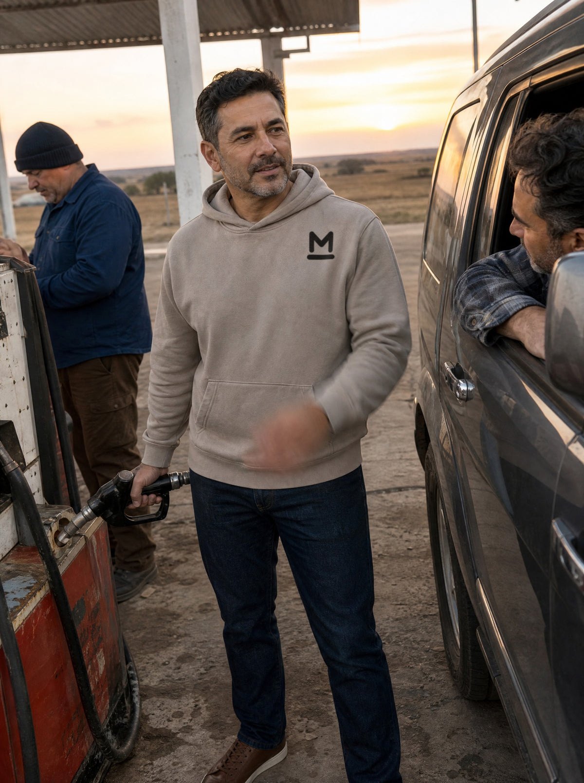 Man in a Mushroom El Camino Hoodie pumping gas at a rural station at sunrise, chatting with a friend