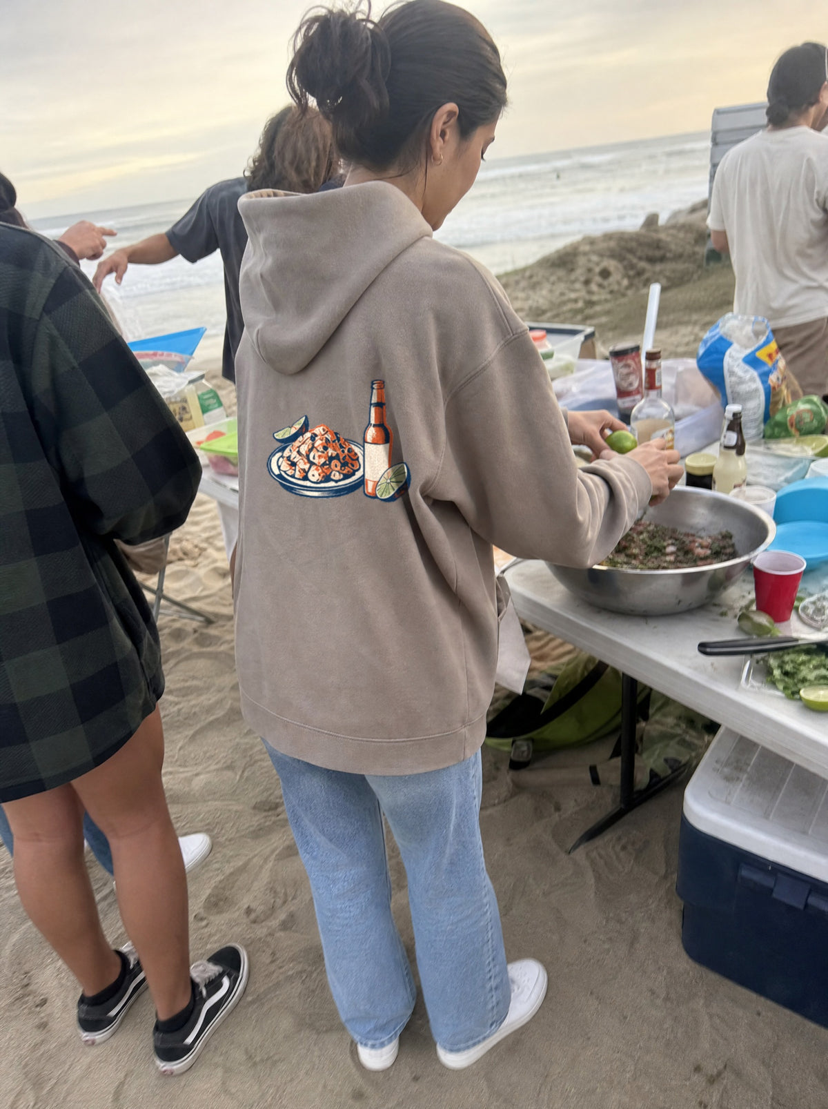 Woman in a Mushroom El Ceviche Hoodie, back view, squeezing limes into a bowl at a beach cookout