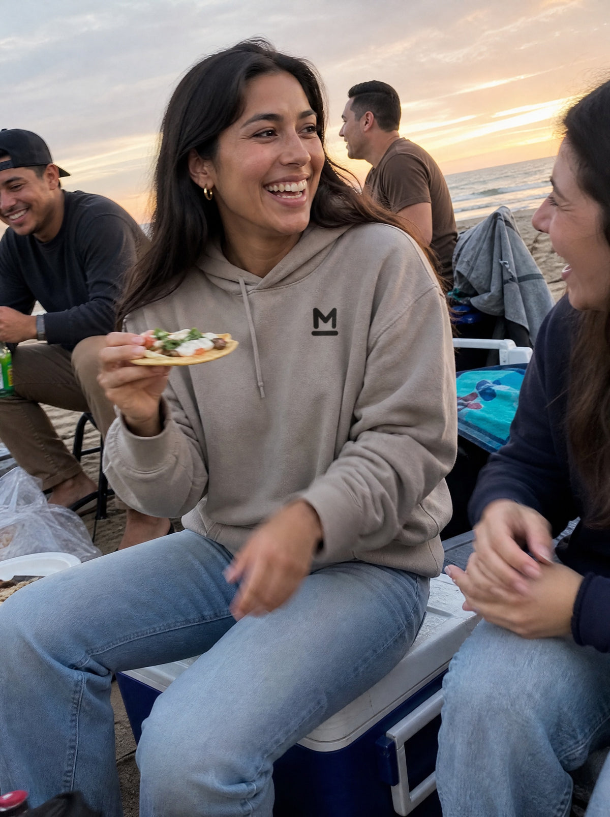Woman in a Mushroom El Ceviche Hoodie eating a tostada on a cooler at a beach cookout