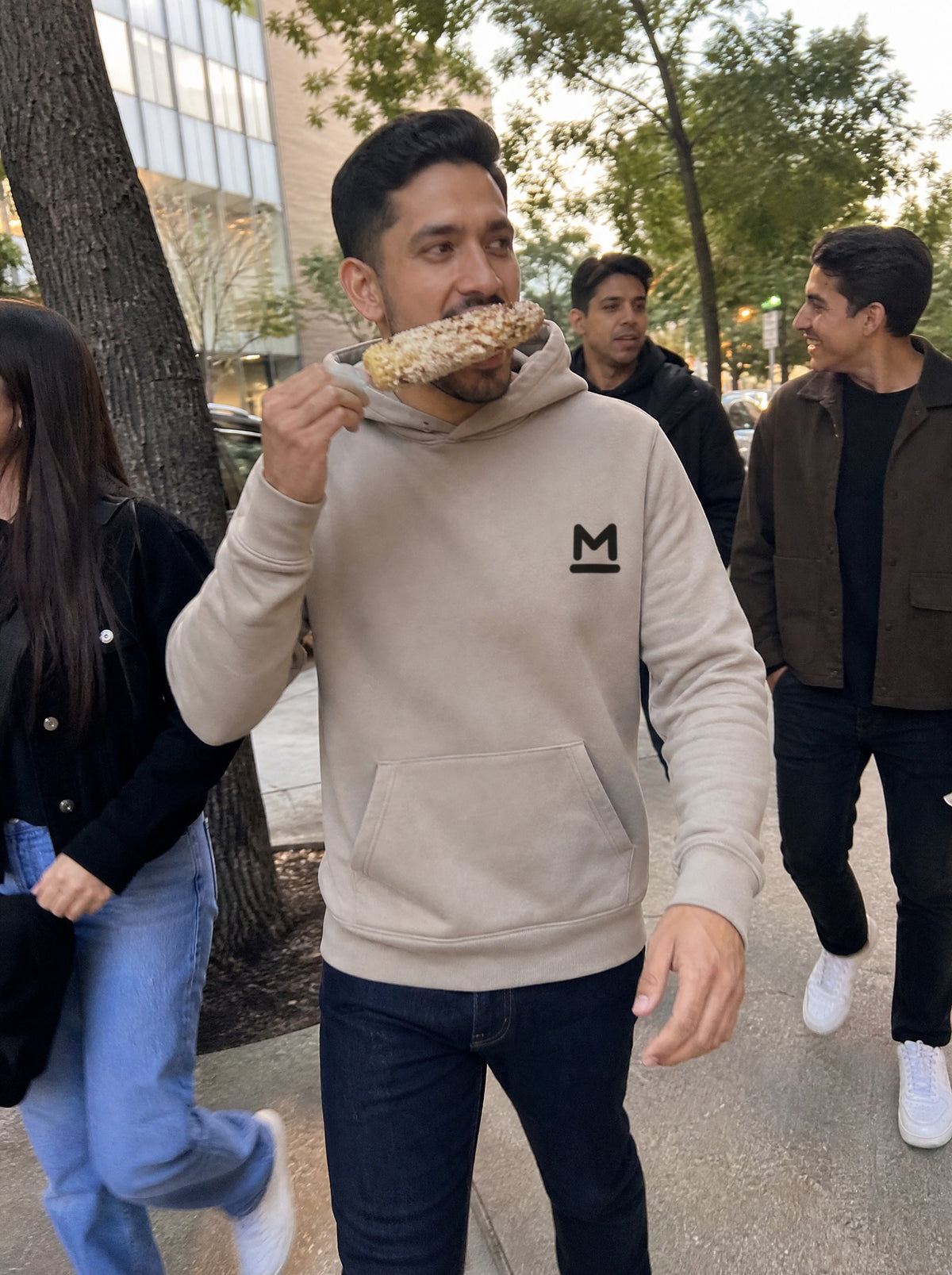 Man in a Mushroom El Elote Hoodie eating an elote on a city sidewalk with friends