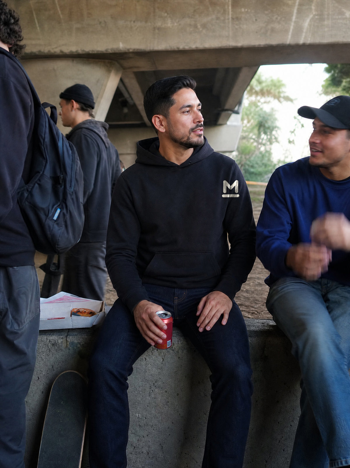 Man in a Black El Viaducto Hoodie sitting on a wall under a freeway bridge with friends