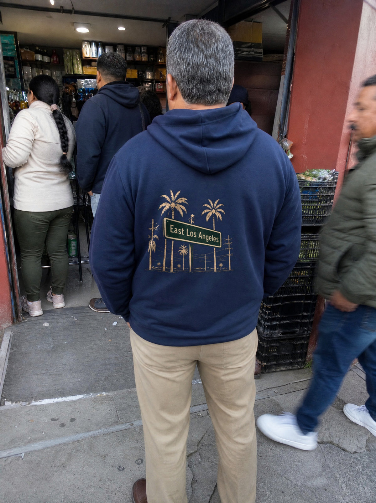 Man in a Navy Blue La Esquina Hoodie, back view, in line outside a corner store
