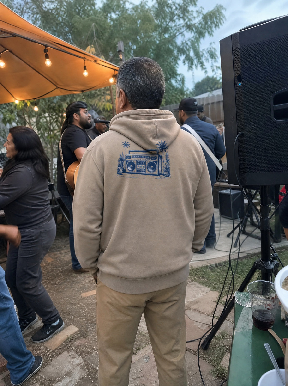 Man in a Mushroom La Música Hoodie, back view, by a speaker and friends with guitars at a party