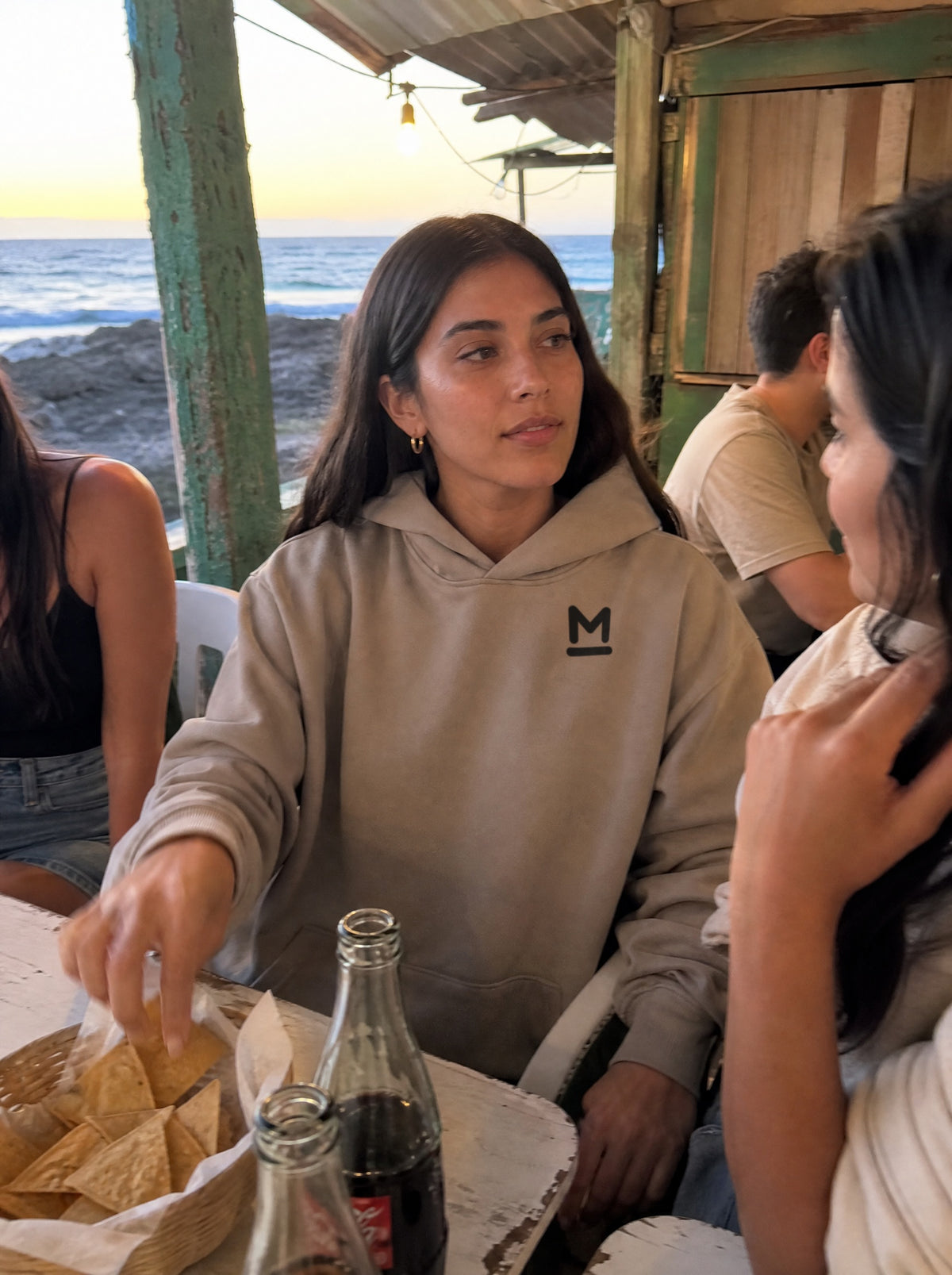Woman in a Mushroom La Palapa Hoodie reaching for chips at a seaside shack with friends
