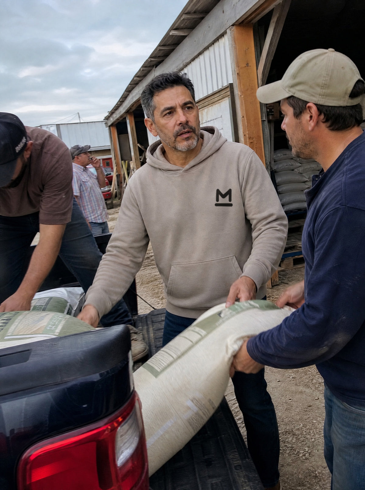 Man in a Mushroom La Reata Hoodie loading feed sacks into a pickup at a rural feed store