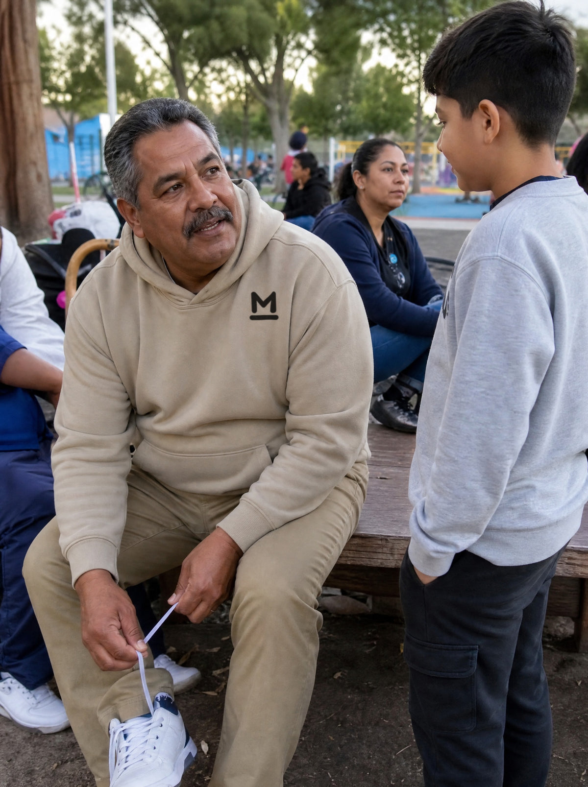 Man in a Mushroom Los Tenis Hoodie lacing up sneakers on a park bench while his grandson waits