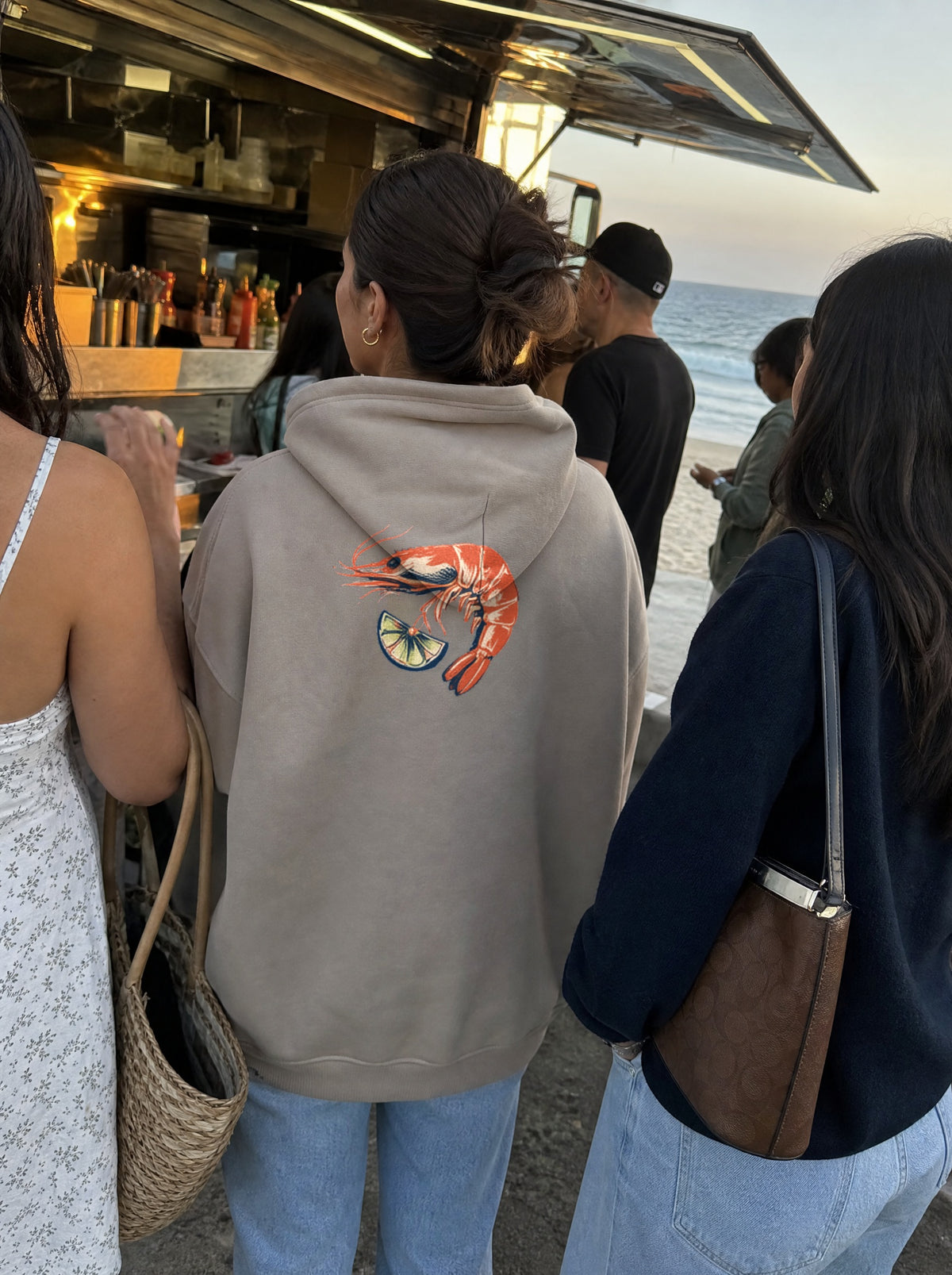 Woman in a Mushroom El Camarón Hoodie, back view, in line at a beachside mariscos truck