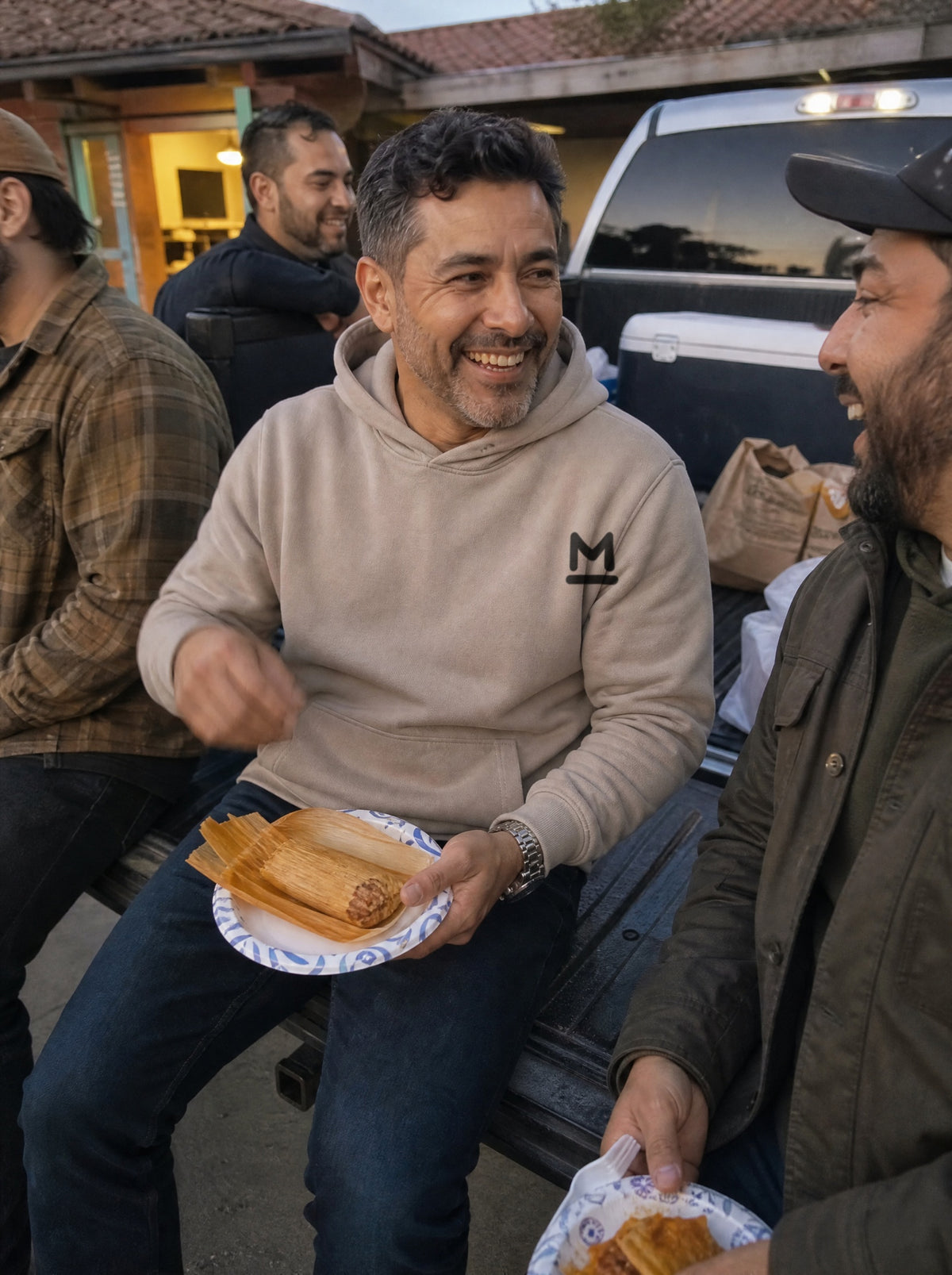 Man in a Mushroom El Corral Hoodie eating a tamal on a truck tailgate at dusk with a friend
