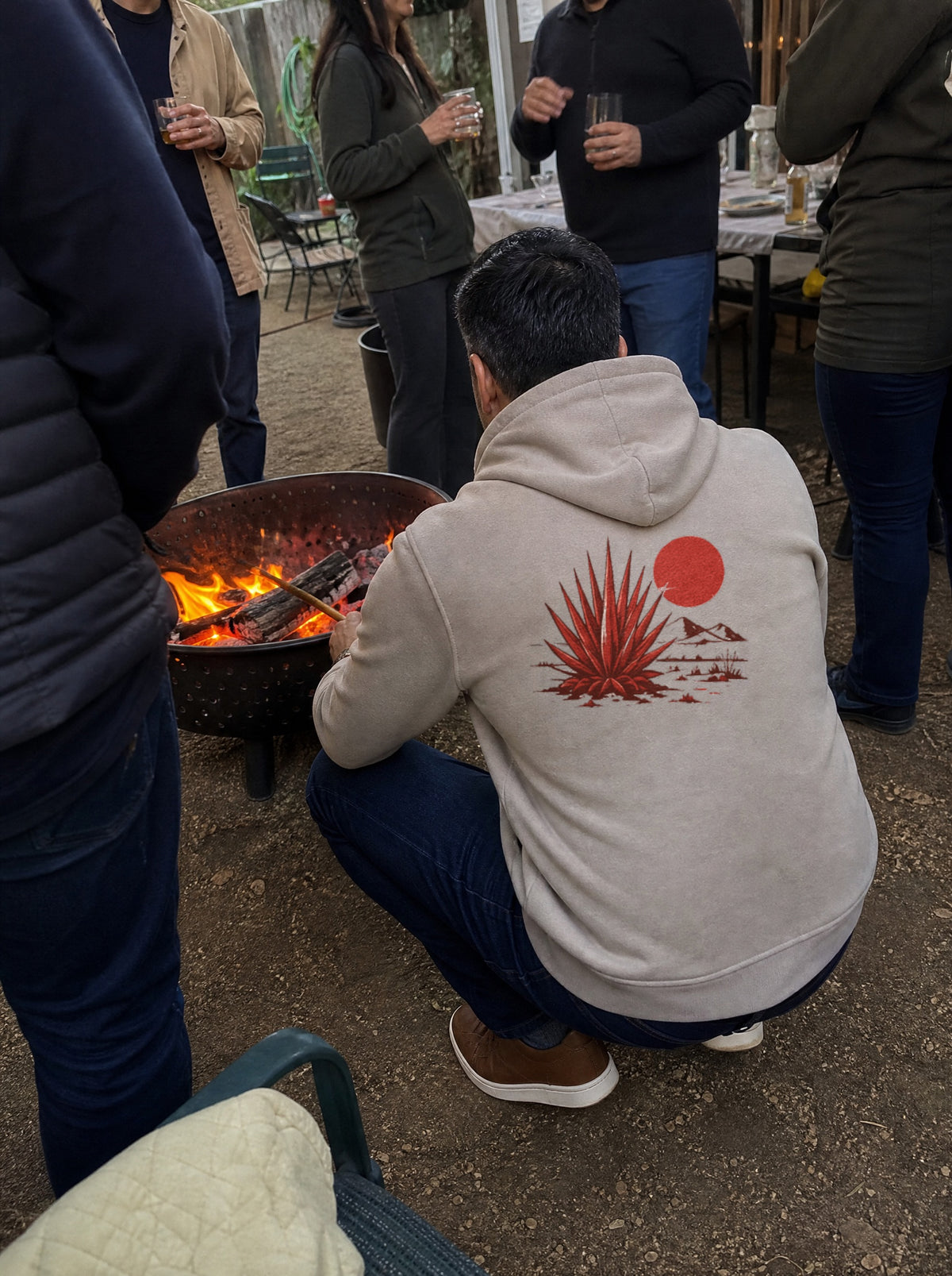 Man in a Mushroom El Mezquite Hoodie, back view, tending a mesquite fire pit with family