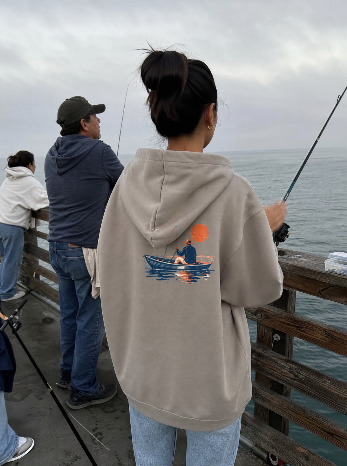 Woman in a Mushroom El Pescador Hoodie, back view, casting a line off a pier with her dad