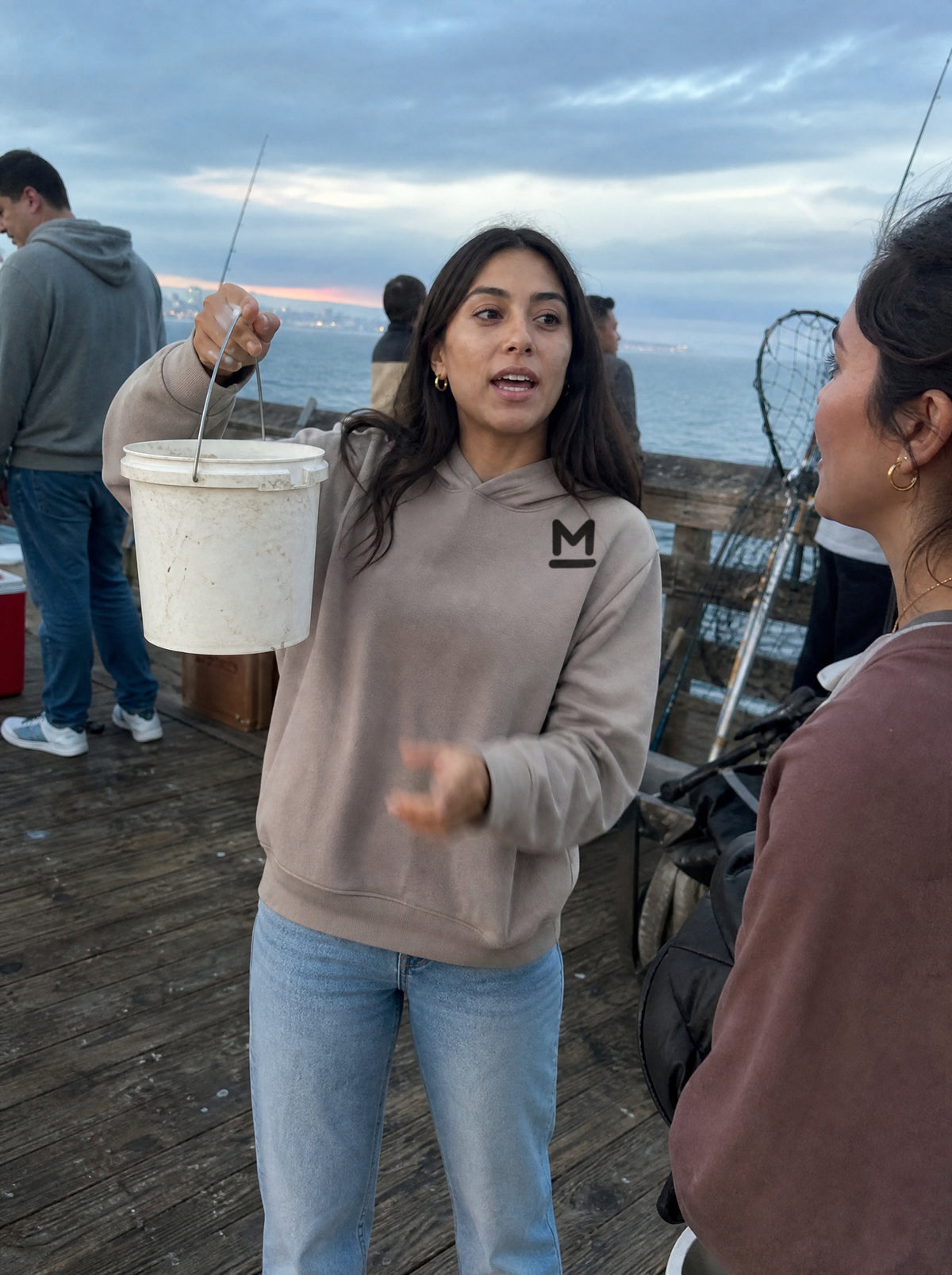 Woman in a Mushroom El Pescador Hoodie holding up a bucket on a fishing pier with her cousin