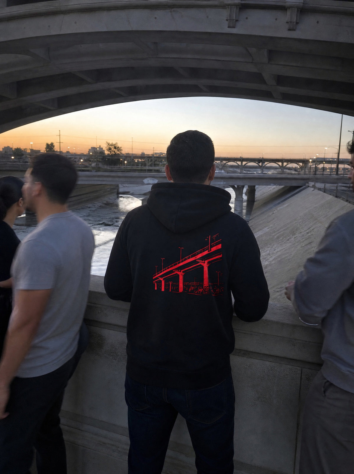 Man in a Black El Viaducto Hoodie, back view, at a river channel overlook under a bridge at dusk