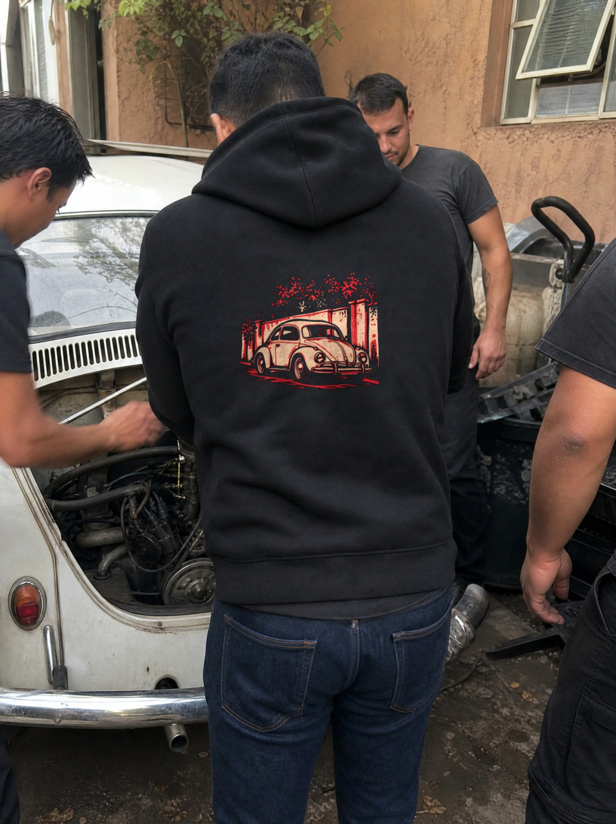 Man in a Black El Vocho Hoodie, back view, working on an old vocho engine with a friend