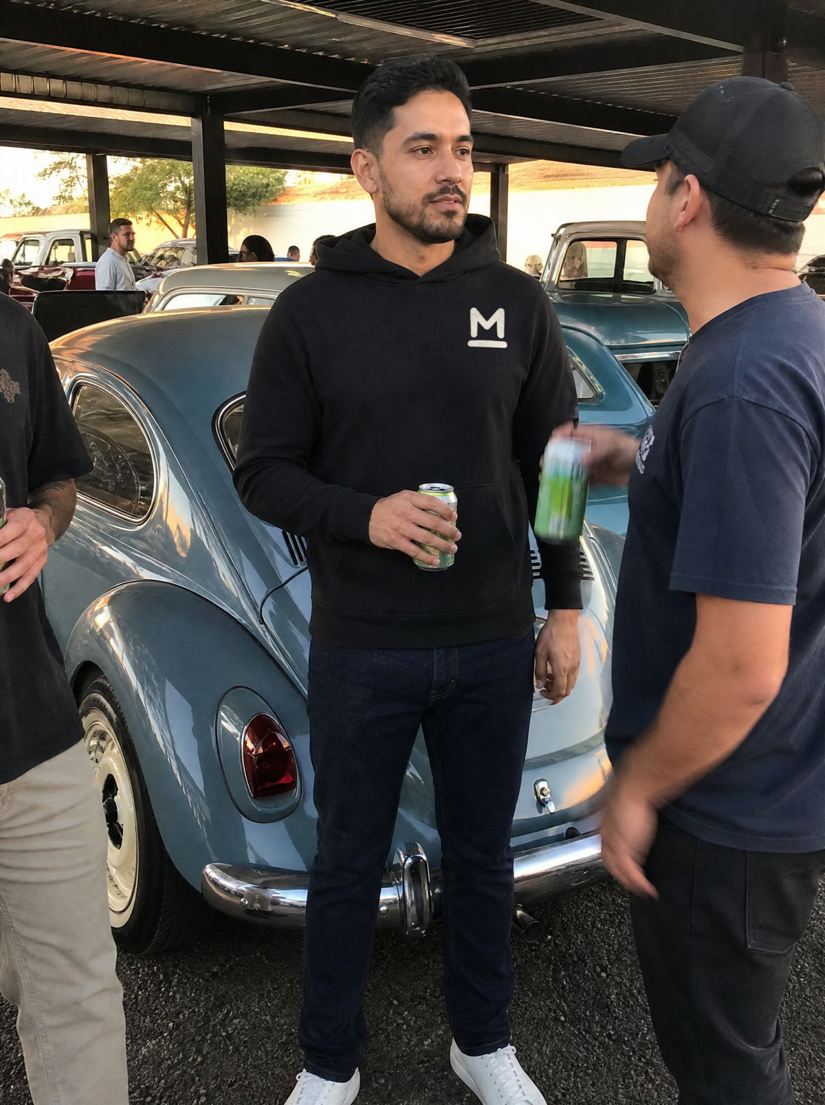 Man in a Black El Vocho Hoodie standing by an old vocho at a car meet with friends