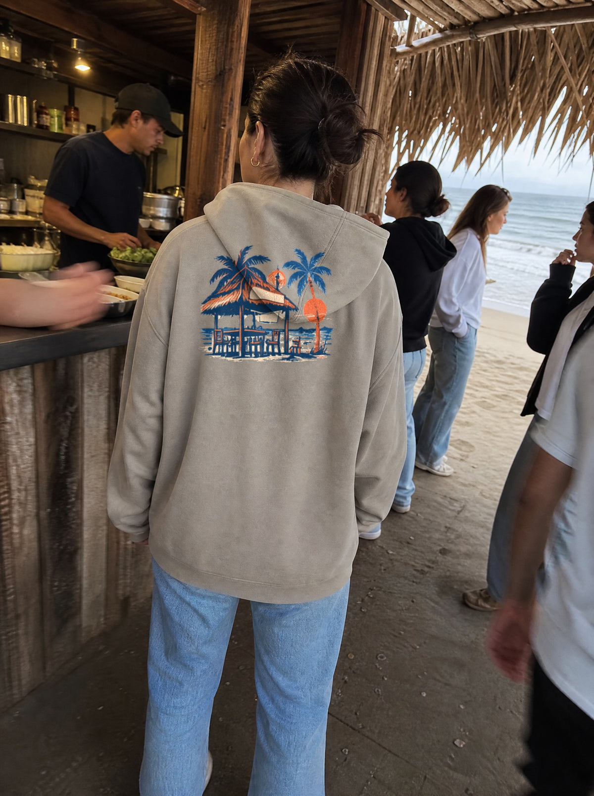 Woman in a Mushroom La Palapa Hoodie, back view, ordering at a seaside palapa food shack